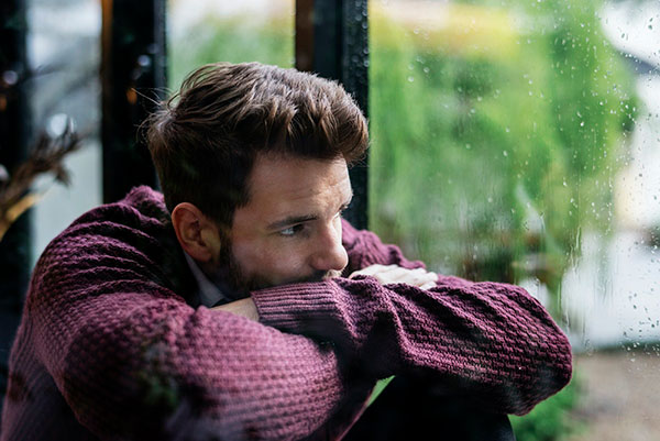 Young adult man sitting by a window looking out at the rain, leaning his head on his hands