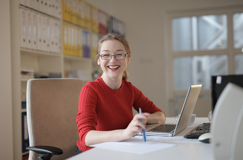 woman smiling, seated with laptop on desk woman smiling, seated with laptop on desk