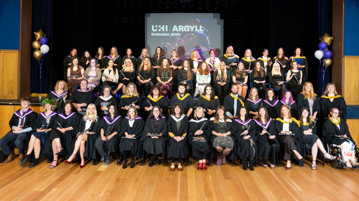 Graduates in gowns pose on stage at the UHI Argyll ceremony, celebrating with graduation scrolls, and balloons. Graduates in gowns pose on stage at the UHI Argyll ceremony, celebrating with graduation scrolls, and balloons.