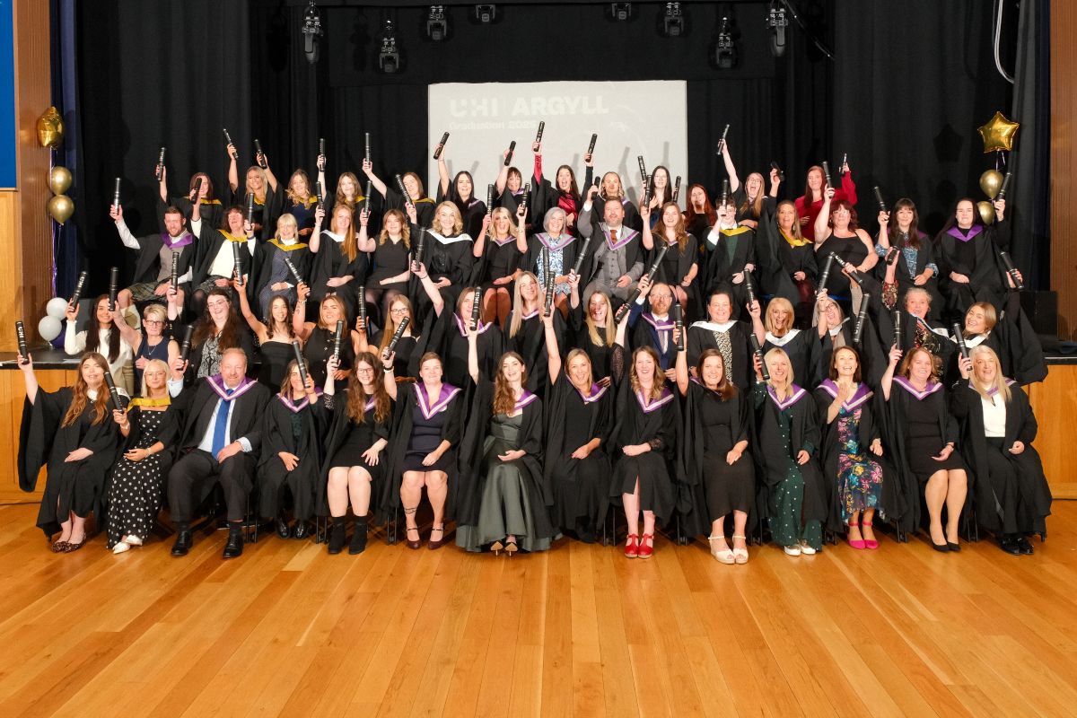 Graduates in gowns pose on stage at the UHI Argyll ceremony, celebrating with graduation scrolls, and balloons.